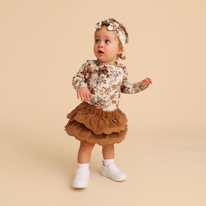 Baby standing, full body, wearing the camel corduroy scalloped bummie with a floral top and headband